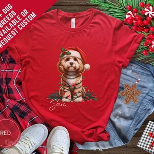 May include: A red t-shirt with a festive illustration of a dog wearing a Santa hat and wrapped in Christmas lights. The shirt has the text "Sam" and "100+ DOG BREEDS AVAILABLE OR REQUEST CUSTOM".