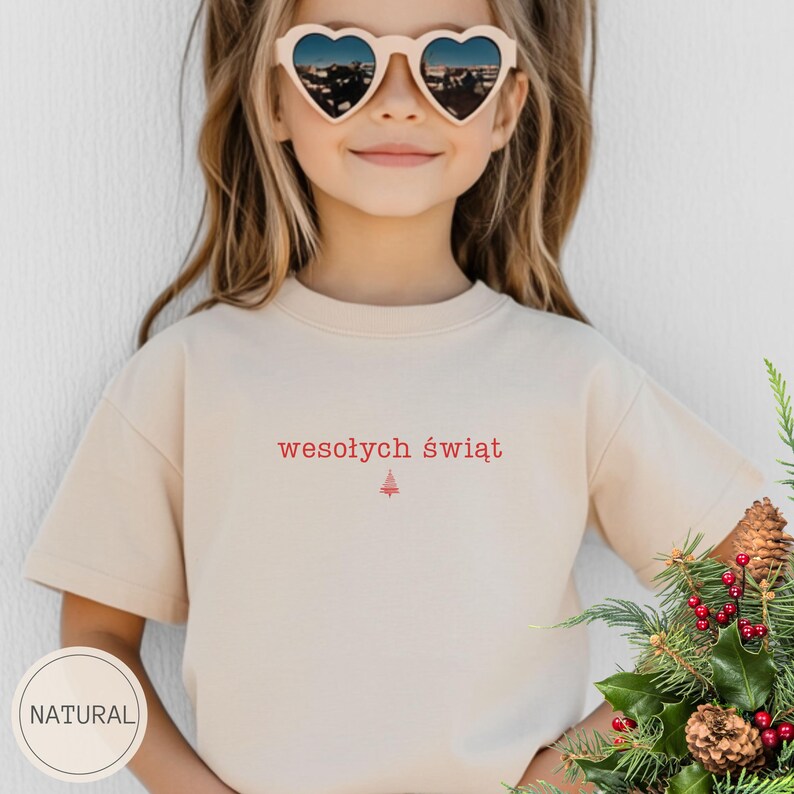 Może przedstawiać: Beżowy t-shirt z kr&oacute;tkim rękawem z czerwonym napisem "wesołych świąt" wyśrodkowanym na klatce piersiowej, umieszczonym nad małą czerwoną grafiką choinki. Koszulka posiada okrągły dekolt i wykonana jest z gładkiego, jednolitego materiału.