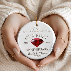 May include: A round white ceramic ornament featuring a red ruby gemstone graphic in the center. The text reads OUR RUBY ANNIVERSARY, 1987, 2027, Amelia & Thomas, and 40 Years. A thin gold string is attached to the top for hanging purposes.