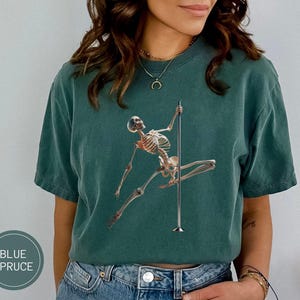 May include: A blue spruce t-shirt features a skeleton figure pole dancing. The graphic is in a realistic style. The shirt is a solid color and has a relaxed fit. The text "BLUE SPRUCE" is in a circle on the bottom left.