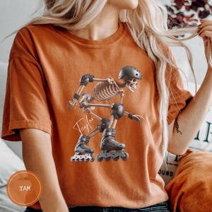 May include: Orange t-shirt featuring a skeleton rollerblading, complete with a helmet and protective gear. The name "Taylor" is written in cursive. The shirt has a round neck and short sleeves.