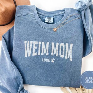 Op de afbeelding: Een spijkerbroekblauwe sweatshirt met de tekst "WEIM MOM" in witte blokletters. Onder de tekst staat de naam "LUNA" en een pootafdruk. Een gouden ketting met een hanger is over de sweatshirt gedrapeerd.