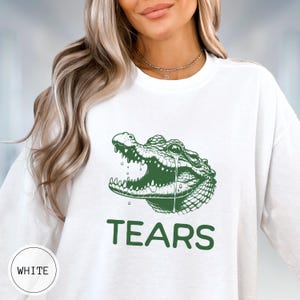 May include: A white long-sleeve sweatshirt featuring a green graphic of an alligator head with an open mouth and a single tear falling from its eye. The word TEARS is printed in large green capital letters directly below the alligator illustration.