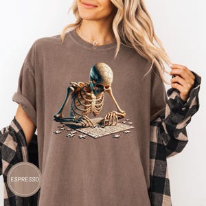May include: Espresso-colored t-shirt featuring a detailed illustration of a skeleton assembling a jigsaw puzzle. The skeleton's skull is a puzzle piece. The shirt has a relaxed fit and a round neckline. The word "ESPRESSO" is printed on the bottom left.