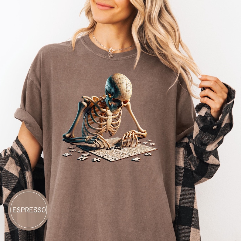 May include: Espresso-colored t-shirt featuring a detailed illustration of a skeleton assembling a jigsaw puzzle. The skeleton's skull is a puzzle piece. The shirt has a relaxed fit and a round neckline. The word "ESPRESSO" is printed on the bottom left.