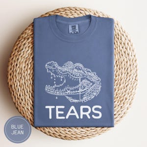 May include: A blue jean colored t-shirt featuring a white line art graphic of an alligator with its mouth open and a single tear falling from its eye. The word TEARS is printed in white capital letters below the alligator graphic on the chest.