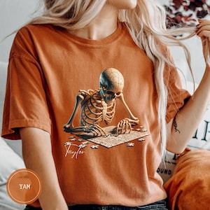 May include: Burnt orange t-shirt featuring a detailed illustration of a skeleton assembling a puzzle. The skeleton's skull is a puzzle piece, and the name "Taylor" is written in cursive below the puzzle. The shirt has a relaxed fit.