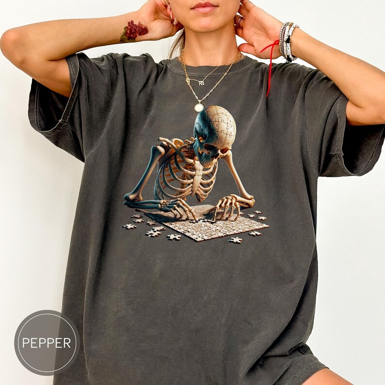 May include: Dark gray t-shirt featuring a detailed graphic of a skeleton assembling a jigsaw puzzle. The skeleton's skull is a puzzle piece, and other pieces are scattered around. The shirt has a relaxed fit.
