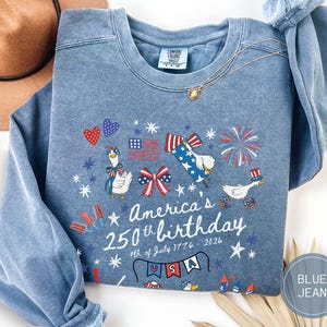May include: Blue jean-coloured sweatshirt with a patriotic design. The graphic includes cartoon geese, fireworks, and the text "America's 250th birthday 4th of July 1776-2026". The design features red, white, and blue elements.
