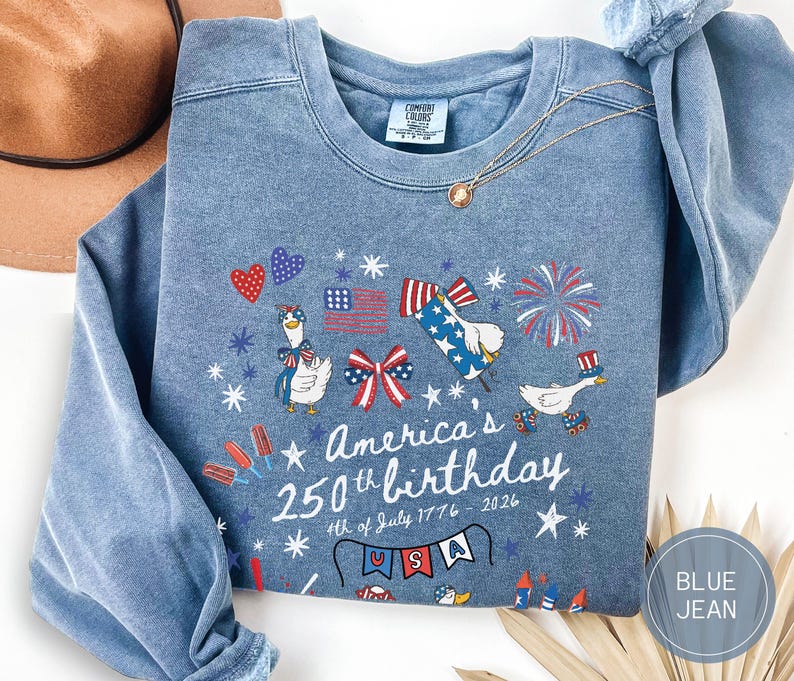 May include: Blue jean-coloured sweatshirt with a patriotic design. The graphic includes cartoon geese, fireworks, and the text "America's 250th birthday 4th of July 1776-2026". The design features red, white, and blue elements.