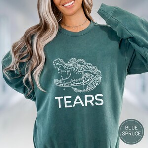 May include: A blue spruce colored crewneck sweatshirt featuring a white line art illustration of an alligator head with an open mouth and the word TEARS printed in white capital letters underneath the graphic on the front of the garment.