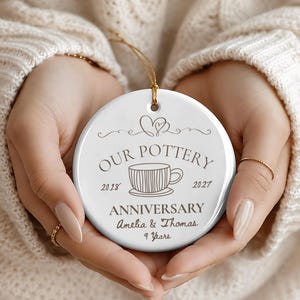 May include: A round white ceramic ornament featuring a gold hanging string. The design includes a line drawing of a teacup, a heart icon, and the text: OUR POTTERY ANNIVERSARY, 2018, 2027, ANNIVERSARY, Amelia & Thomas, and 9 Years in dark brown ink.