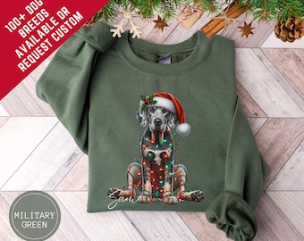 Op de afbeelding: Militairgroen sweatshirt met een zittende hond met een kerstmuts en kerstverlichting. De hond heeft een zwart-witte gevlekte vacht. Tekst linksboven luidt "100+ DOG BREEDS AVAILABLE OR REQUEST CUSTOM".