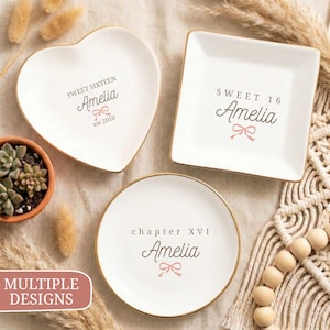 Könnte beinhalten: Drei weiße Keramikplatten mit goldenen Rändern, jede in einer anderen Form: Herz, Quadrat und rund. Jede Platte trägt den Namen "Amelia" und eine Schleifengrafik mit Text, der ein Sweet Sixteen- oder Kapitel XVI-Thema angibt.