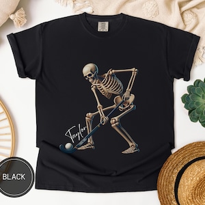 以下が含まれることがあります： フィールドホッケーをしているスケルトンのデザインがプリントされた黒いTシャツ。スケルトンはスティックとボールを持っています。Tシャツには「テイラー」という文字が筆記体で書かれています。
