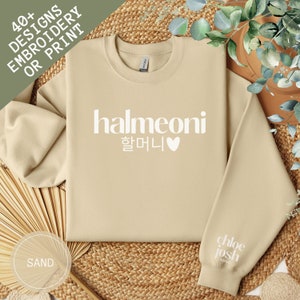 May include: A sand-colored sweatshirt with the text "halmeoni 할머니" and a heart in white. The sweatshirt has a white cuff with the text "chloe josh" in black.