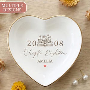 May include: A white heart-shaped ceramic trinket dish with a gold-toned rim. The center features a brown graphic of an open book with flowers, the numbers 20 and 08, the text "Chapter Eighteen", the name "AMELIA", and a small red heart icon.