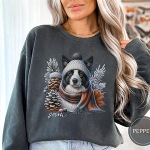 May include: A dark gray sweatshirt with a black and white border collie wearing a gray and white knit hat and a red and black scarf. The dog is surrounded by pine cones and greenery. The text "Sam" is printed below the dog.