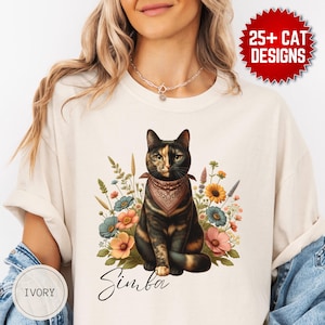 Può includere: T-shirt color avorio con un disegno di gatto. La grafica mostra un gatto con una bandana, circondato da fiori, con il nome "Simba" sotto. Il testo "25+ CAT DESIGNS" è anche visibile.