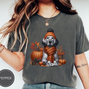 May include: A gray t-shirt with a graphic of a dog wearing a brown and orange hat and scarf. The dog is surrounded by fall-themed elements like pumpkins, leaves, and berries. The text "Pepper" is visible in the lower left corner of the image.