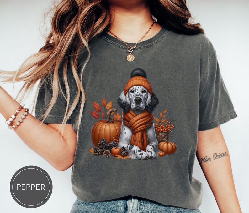 May include: A gray t-shirt with a graphic of a dog wearing a brown and orange hat and scarf. The dog is surrounded by fall-themed elements like pumpkins, leaves, and berries. The text "Pepper" is visible in the lower left corner of the image.