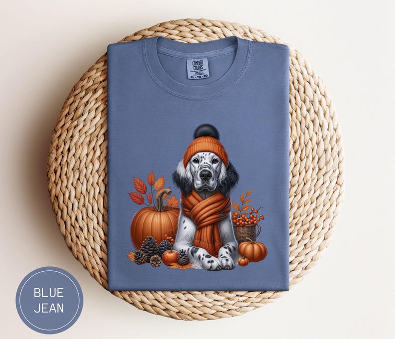 May include: A blue jean t-shirt with a white and black dog wearing a brown and orange knitted hat and scarf. The dog is surrounded by fall foliage, pumpkins, and acorns.