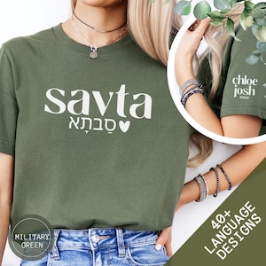 May include: Military green t-shirt with the word "Savta" in large white letters, along with Hebrew text and a heart symbol. The sleeve features the text "chloe josh xoxo". The shirt is paired with blue jeans.