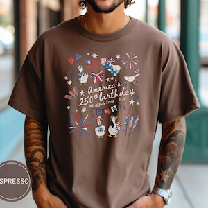 May include: Espresso-coloured t-shirt featuring a graphic commemorating America's 250th birthday. The design includes fireworks, flags, and cartoon geese in patriotic attire. The text reads "America's 250th birthday 4th of July 1776 - 2026."