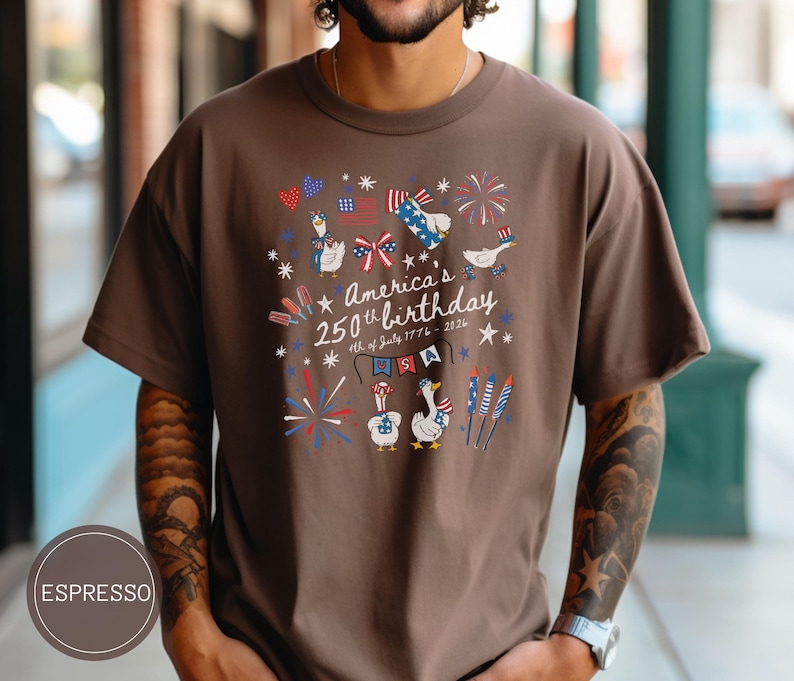 May include: Espresso-coloured t-shirt featuring a graphic commemorating America's 250th birthday. The design includes fireworks, flags, and cartoon geese in patriotic attire. The text reads "America's 250th birthday 4th of July 1776 - 2026."