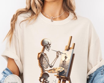 May include: Ivory-colored t-shirt featuring a skeleton artist painting on a canvas. The skeleton holds a paintbrush and palette, with the artwork depicting another skeleton. The artist's signature "Taylor" is visible. The shirt is a casual, comfortable style.