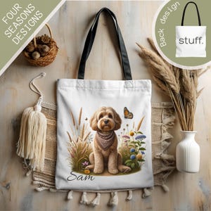 May include: White tote bag featuring a watercolor illustration of a dog wearing a bandana, surrounded by wildflowers and tall grass. The dog's name, "Sam," is written below. The bag has black handles and the back design says "stuff."