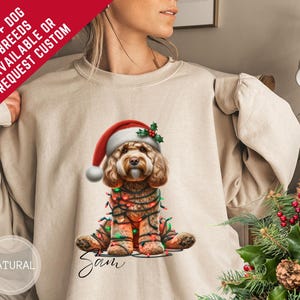 Peut inclure: Un sweat-shirt beige avec un motif de chien de Noël. Le chien, orné d'un bonnet de Père Noël et de guirlandes lumineuses, est l'élément central. Le sweat-shirt porte également le texte "100+ DOG BREEDS AVAILABLE OR REQUEST CUSTOM" et le nom "Sam".