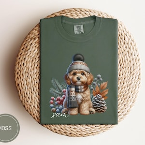 May include: A green t-shirt featuring a charming illustration of a dog wearing a winter hat and scarf. The dog is surrounded by berries and leaves. The text "Sam" is printed below the dog.