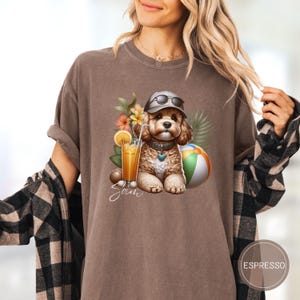 May include: A brown t-shirt with a graphic of a dog wearing sunglasses and holding a beach ball. The dog is surrounded by tropical flowers and a glass of orange juice. The text "Sam" is printed below the graphic.