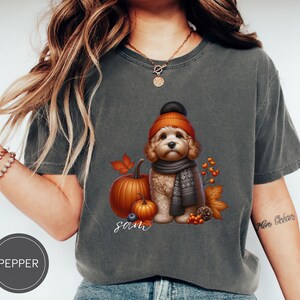 Peut inclure: Un t-shirt gris avec un motif représentant un chien portant un chapeau marron et orange et une écharpe. Le chien est entouré de feuilles d'automne, de citrouilles et de baies. Le texte "Sam" est imprimé sous le chien.