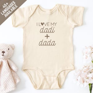 Pode incluir: Body de bebé creme com o texto "I love my dadi + dada" em castanho. Um ursinho de pelúcia e flores estão no fundo. O body é de manga curta e tem um design simples, adequado para um bebé.