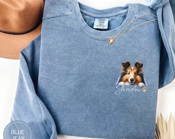 Op de afbeelding: Blauwe jeans sweatshirt met een afbeelding van een Shetland Sheepdog die uit een gat kijkt met de tekst "Shadow" eronder.