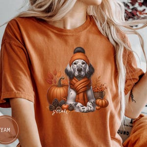 May include: Orange t-shirt with a white and brown dog wearing a brown and orange hat and scarf. The dog is surrounded by fall-themed elements like pumpkins, leaves, and berries. The text "Sam" is printed on the shirt.