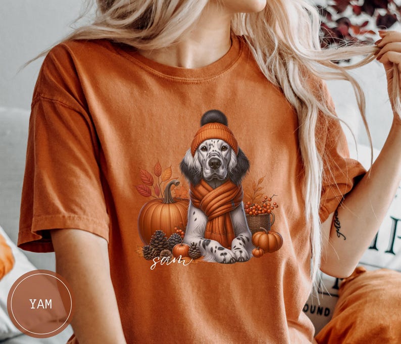 May include: Orange t-shirt with a white and brown dog wearing a brown and orange hat and scarf. The dog is surrounded by fall-themed elements like pumpkins, leaves, and berries. The text "Sam" is printed on the shirt.