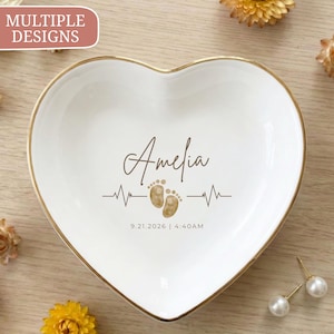 May include: A white heart-shaped ceramic trinket dish with a gold-toned rim. The center features the name Amelia in brown script, a brown heartbeat line graphic, two small baby footprints, and the date 9.21.2026 with the time 4:40AM.