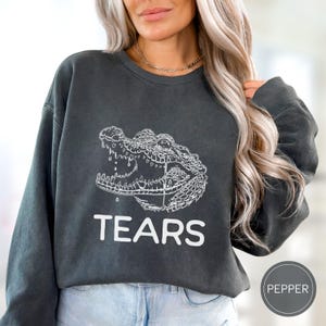 May include: A charcoal gray crewneck sweatshirt featuring a white line art illustration of an alligator head with its mouth open and a single tear falling from its eye. The word TEARS is printed in white capital letters below the alligator graphic.