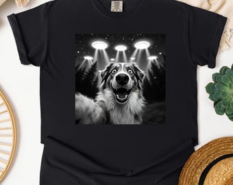 May include: A black and white graphic t-shirt featuring a dog with its mouth open and tongue out, looking up at a group of UFOs in the sky. The dog is in a forest setting.