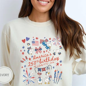 May include: Ivory long-sleeved shirt featuring a design celebrating America's 250th birthday. The design includes fireworks, flags, stars, and cartoon geese wearing patriotic accessories. The text reads "America's 250th birthday 4th of July 1776-2026".