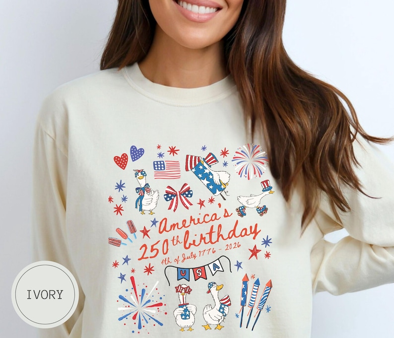 May include: Ivory long-sleeved shirt featuring a design celebrating America's 250th birthday. The design includes fireworks, flags, stars, and cartoon geese wearing patriotic accessories. The text reads "America's 250th birthday 4th of July 1776-2026".