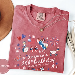May include: Crimson t-shirt featuring a patriotic design for America's 250th birthday. The graphic includes fireworks, flags, and cartoon geese. The text reads "America's 250th birthday 4th of July 1776-2026."