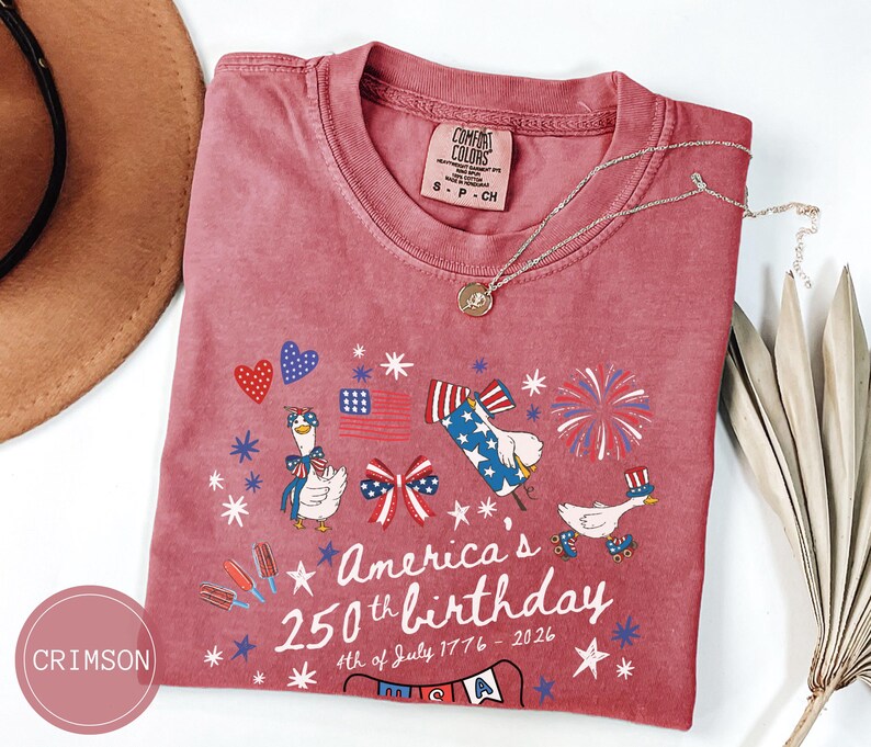 May include: Crimson t-shirt featuring a patriotic design for America's 250th birthday. The graphic includes fireworks, flags, and cartoon geese. The text reads "America's 250th birthday 4th of July 1776-2026."