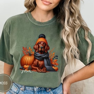 May include: A green t-shirt with a graphic of a dog wearing a hat and scarf with pumpkins and fall leaves. The text "Sam" is printed on the shirt.