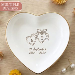 Puede incluir: Un plato de cerámica blanco en forma de corazón con un borde dorado. El centro presenta un dibujo lineal marrón de dos relicarios en forma de corazón unidos por un lazo, con las letras A y T, y el texto 21 September 2017 - 2027 debajo.