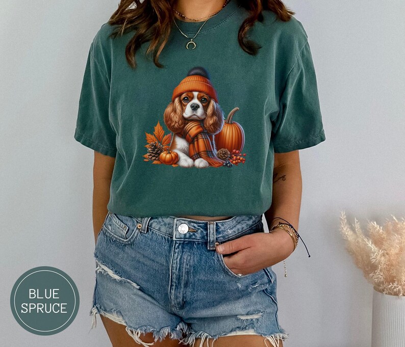 May include: A green t-shirt with a cartoon image of a Cavalier King Charles Spaniel wearing a brown and orange hat and scarf, surrounded by pumpkins and fall leaves.