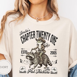 May include: Ivory t-shirt with a graphic of a frog in a cowboy hat riding a raccoon. Text reads "Amelia's Chapter Twenty One EST. 2005 The Plot Thickens."